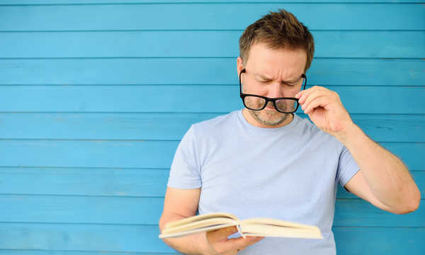 Portret dojrzałego mężczyzny w dużych czarnych okularach, próbującego czytać książkę, ale mającego trudności z widzeniem tekstu z powodu problemów ze wzrokiem