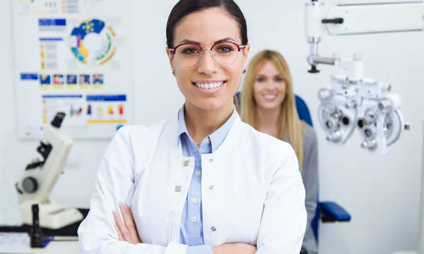 Portret pięknej uśmiechniętej kobiety patrzącej w kamerę w klinice okulistycznej z pacjentką siedzącą na badaniu wzroku